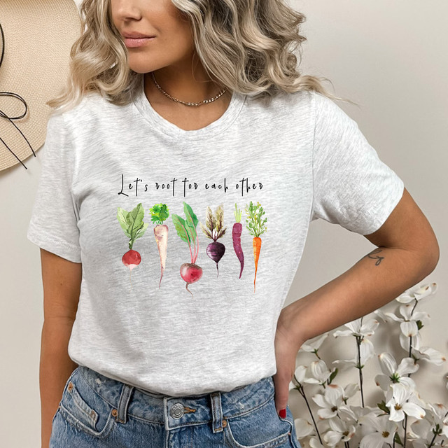Let's Root For Each Other DTF Heat Transfer, image of a smiling woman wearing a light gray T-shirt with a colorful vegetable-themed design and the phrase 'Let's root for each other' printed on it.
