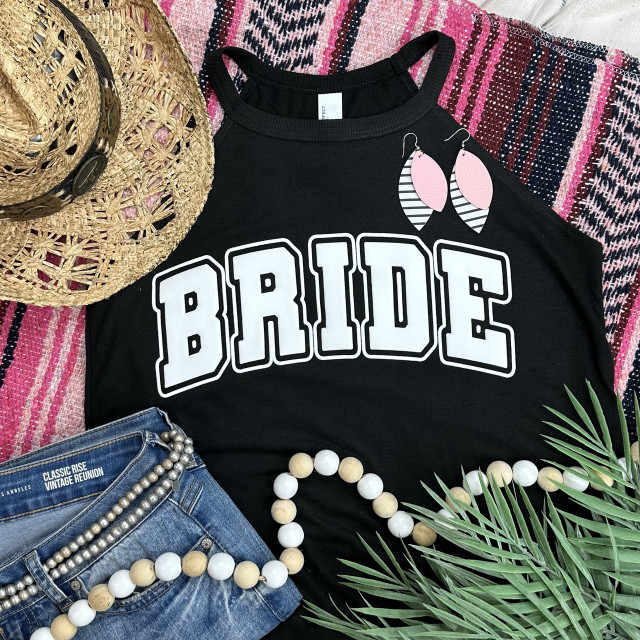 Bride White PUFF Screen Print Heat Transfer, black sleeveless top with 'BRIDE' text design and pink and black striped leaf earrings in a flat lay with denim shorts, straw hat, pearl necklace, and tropical leaves on a pink patterned textile background.