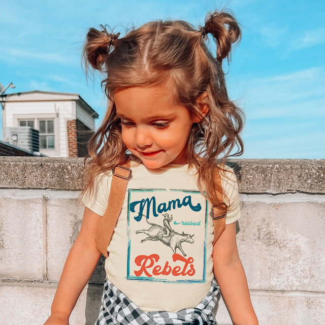Mama Raised Rebels DTF Heat Transfer, a young girl wearing a white T-shirt with a graphic design featuring the words 'Mama Raised Rebels' and a drawing of a playful dog.
