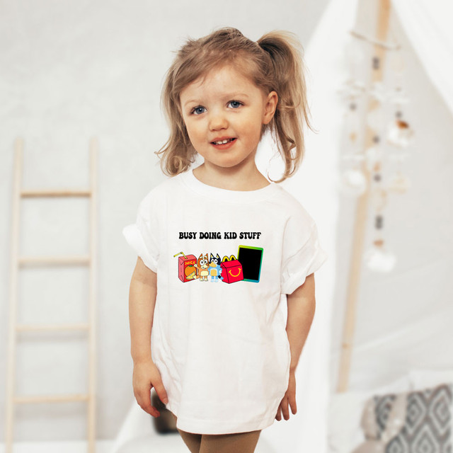 Busy Doing Kid Stuff Blue Dog DTF Heat Transfer, a colorful cartoon graphic of a blue dog, juice box, and tablet on a white T-shirt worn by a young girl.