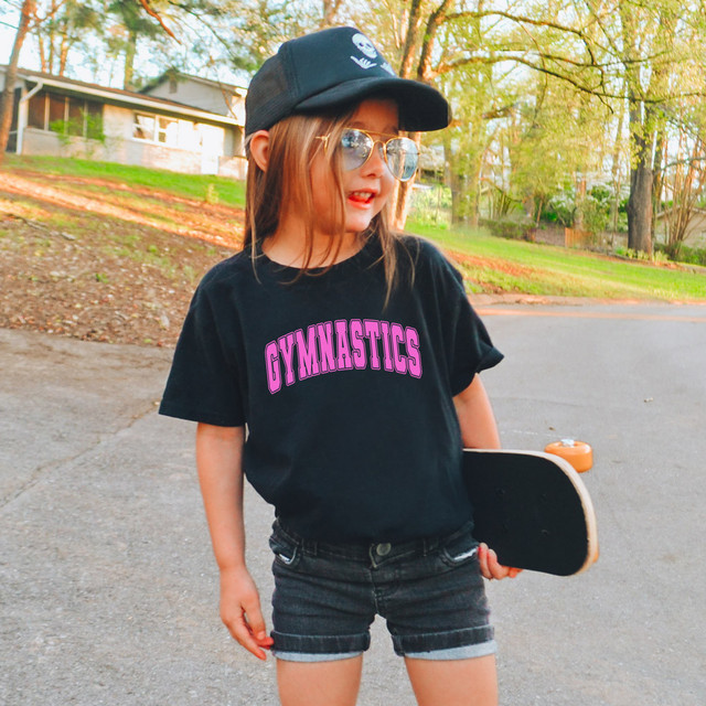 Gymnastics DTF Heat Transfer, girl wearing black t-shirt with pink 'GYMNASTICS' print and holding skateboard outdoors.