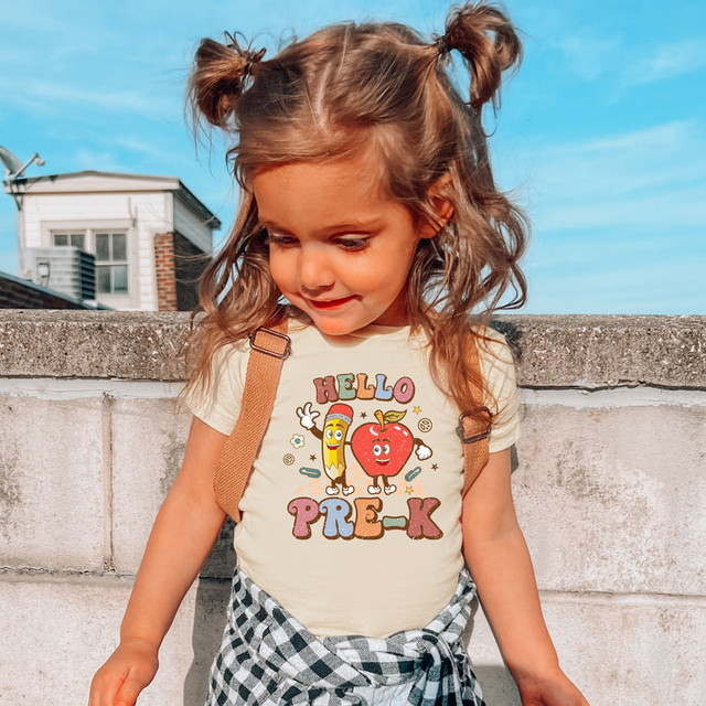 Hello Pre-K Retro DTF Heat Transfer, a colorful design on a young girl's t-shirt featuring animated pencil and apple characters with the text 'Hello Pre-K' on a sunny outdoor day.