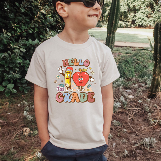 Hello 1st Grade Retro DTF Heat Transfer, a colorful heat transfer design featuring a cheerful pencil and apple character with the text "Hello 1st Grade" on a child's t-shirt.