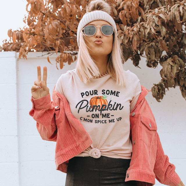 Pour Some Pumpkin On Me C'Mon Spice Me Up DTF Heat Transfer, a woman wearing sunglasses, a beanie, a graphic t-shirt, and a pink jacket, holding up a peace sign outdoors with autumnal leaves in the background.