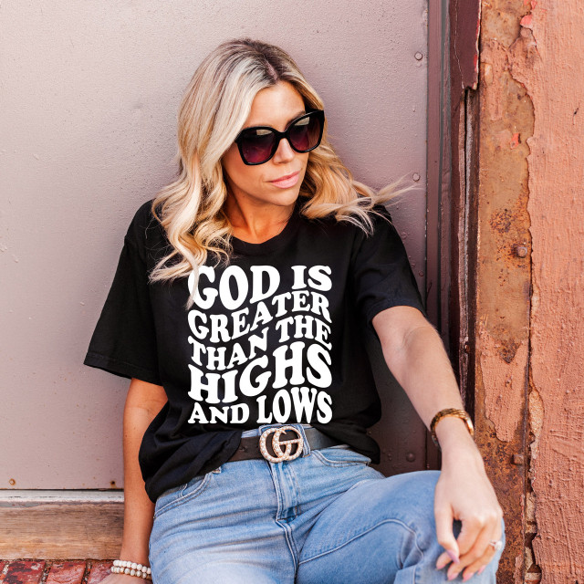 God Is Greater Than The Highs and Lows White DTF Heat Transfer, a woman wearing a black T-shirt with the design, sitting against a textured wall with sunglasses and casual jeans.