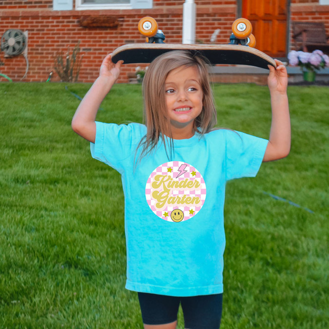 Kindergarten Checkered DTF Heat Transfer, a colorful and playful design featuring a smiling face, stars, and the text 'Kinder Garten' on a girl's blue t-shirt.