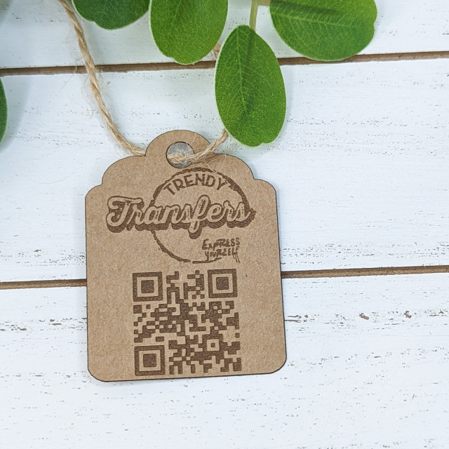Custom Cardstock Hang Tags, a brown kraft paper tag with printed QR code and trendy design on a white wooden surface with green leaves.