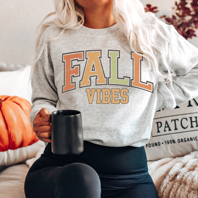 Fall Vibes Pumpkin Screen Print Heat Transfer, woman wearing a cozy sweatshirt with fall-themed text, sitting on a couch with pumpkins and autumn decor.
