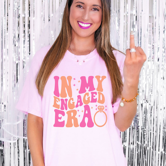 In My Engaged Era DTF Heat Transfer, pink and orange decorative lettering on a light pink T-shirt, with a diamond ring graphic, worn by a woman celebrating her engagement.