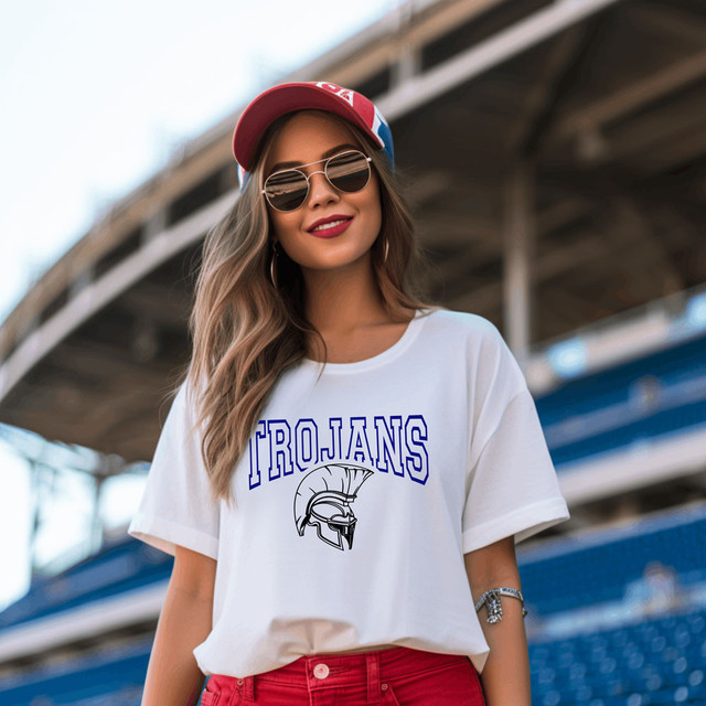 Trojans *CUSTOM COLOR OPTIONS* DTF Heat Transfer, a woman wearing a white T-shirt with Trojans logo and mascot, accessorized with sunglasses, a cap, and jewelry, standing outdoors under a bridge.