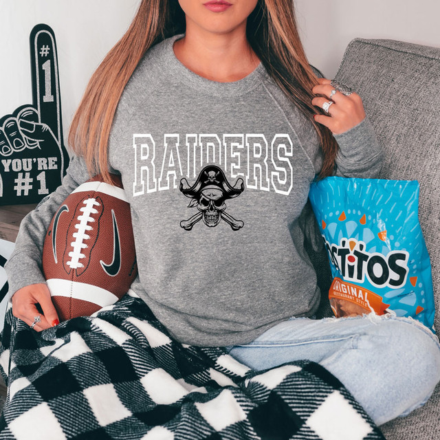 Raiders *CUSTOM COLOR OPTIONS* DTF Heat Transfer, product image of a person wearing a gray sweatshirt with a Raiders logo and skull design, holding a football, sitting next to a bag of Fritos on a gray couch.