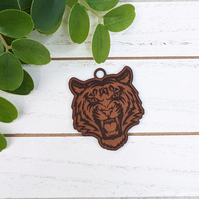 Tiger Mascot Leather Keychain, detailed brown leather keychain featuring a roaring tiger mascot design with black accents on a white wooden surface with green leaves nearby.