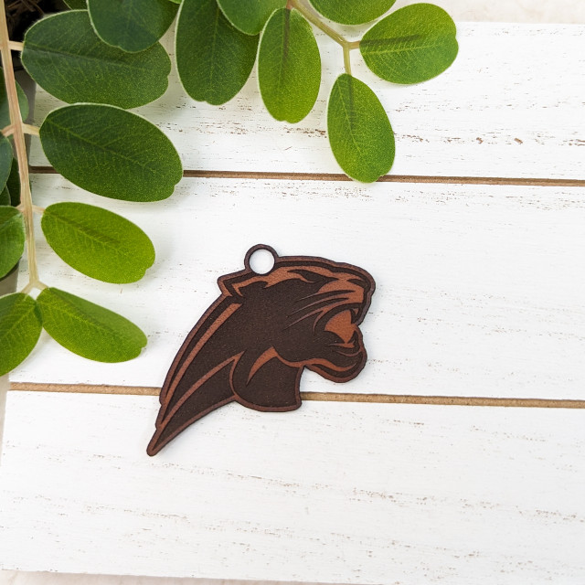 Panther Mascot Leather Keychain, brown and black leather keychain with a panther mascot design on a white wooden surface, surrounded by green leaves.