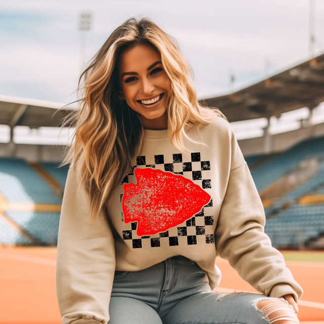 Arrowhead DTF Heat Transfer, a woman wearing a beige sweatshirt with red and black patterned design on the front, sitting outdoors in a stadium.