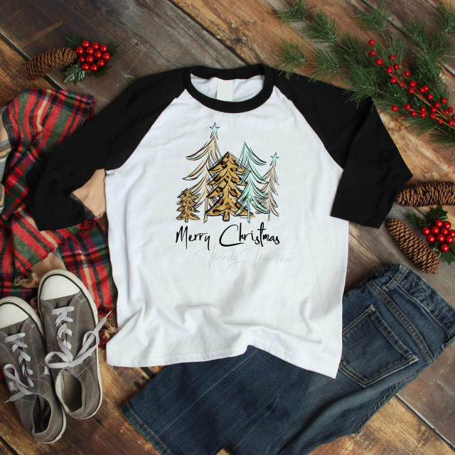 Merry Christmas Trees Sublimation, a graphic tee with hand-drawn style Christmas trees in gold, silver, and teal colors and the text 'Merry Christmas' on a white shirt with black sleeves, displayed with festive holiday decorations.