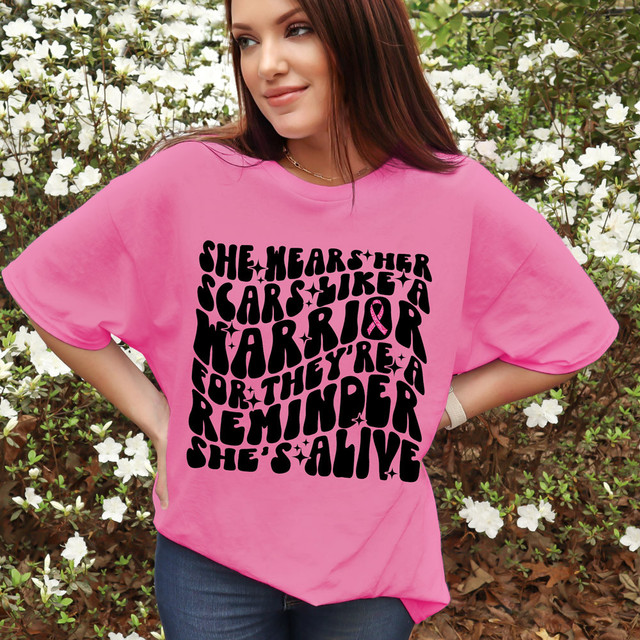 She Wears Her Scars Like A Warrior DTF Heat Transfer, front view of a woman wearing a pink T-shirt with bold black text and pink ribbon design, outdoor setting with white flowers.
