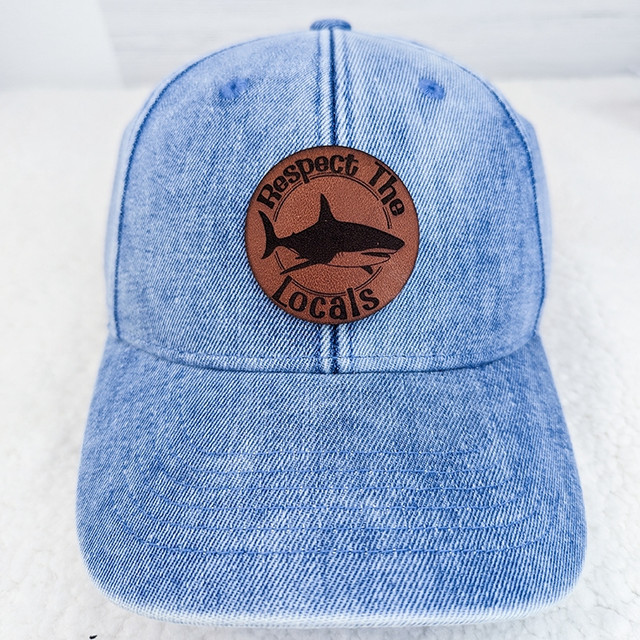 Respect The Locals Leather Hat Patch, showcasing a brown leather patch with a black shark silhouette and the text 'Respect The Locals' embroidered on a light blue denim baseball cap.