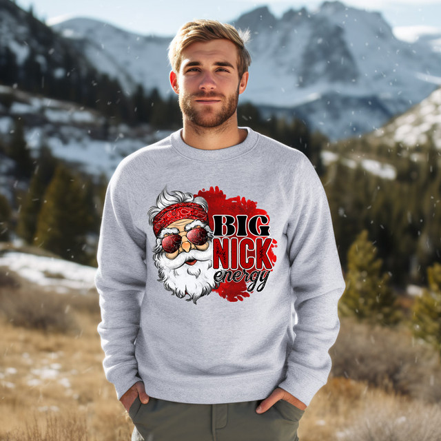 Big Nick Energy DTF Heat Transfer, a creative and vibrant graphic design featuring Santa Claus with sunglasses and a red bandana, placed on a light gray sweatshirt worn by a man outdoors with snow-capped mountains in the background.