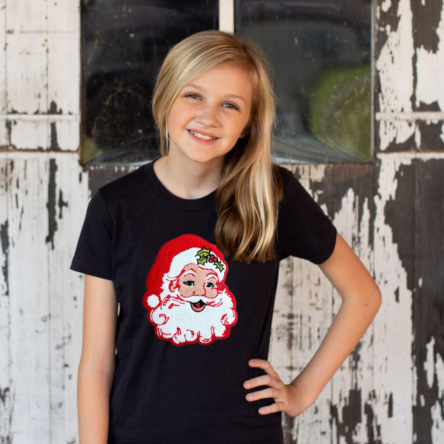 YOUTH LIGHT SKIN Red Santa Chenille Patch, a girl wearing a black T-shirt with a festive Santa Claus patch embroidered on it, standing in front of a rustic wooden backdrop.
