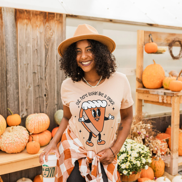 Out Here Lookin' Like A Snack Plain Pie DTF Heat Transfer, woman wearing a graphic t-shirt with a cartoon snack pie and text, smiling at pumpkin patch or fall market.