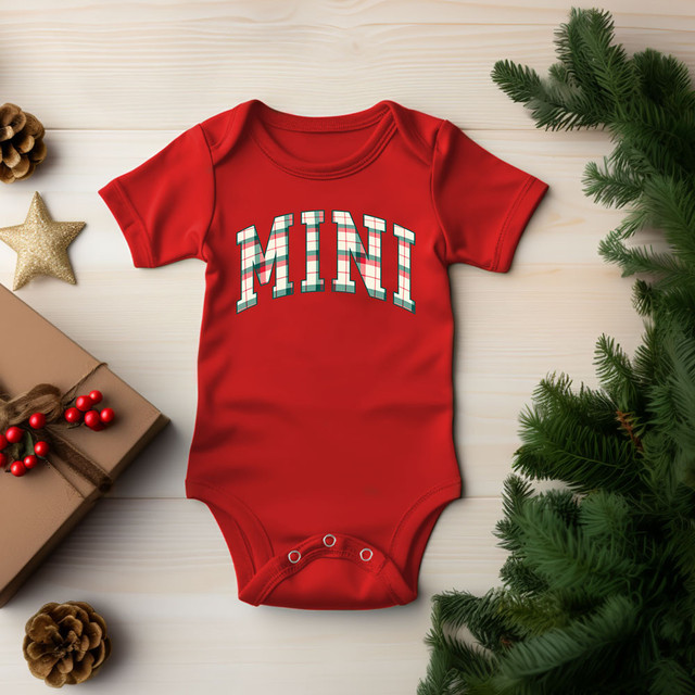 Mini Plaid DTF Heat Transfer, red baby onesie with plaid pattern text 'MINI' on front, holiday-themed background with pine branches, gift box, star and pinecones.