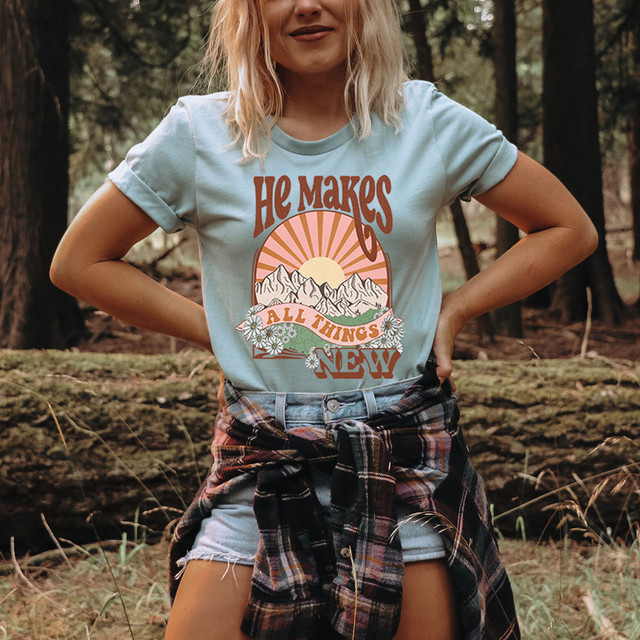He Makes All Things New DTF Heat Transfer, a colorful graphic design of mountains, sun, and flowers on a woman's t-shirt, in an outdoor forest setting.