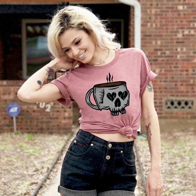 Skeleton Coffee Cup DTF Heat Transfer, illustrated skeleton coffee mug with heart and skull designs on the front of a woman wearing a pink tied-up t-shirt outdoors.