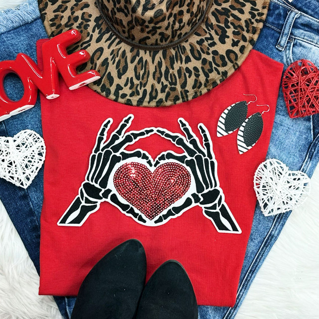 Skeleton Hands + Heart Embroidered Sequin Patch, fashion flat lay with red t-shirt featuring skeleton hands forming a heart with red sequins, surrounded by decorative hearts, a leopard print hat, a pair of black shoes, denim jeans, and red and white woven heart ornaments.