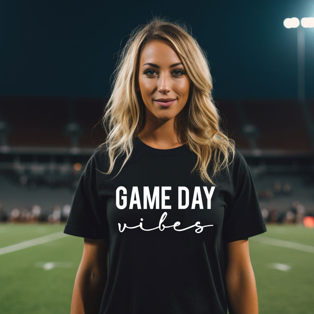 Game Day Vibes WHITE DTF Heat Transfer, a woman wearing a black t-shirt with 'GAME DAY vibes' text printed on it, standing on a sports field at night.