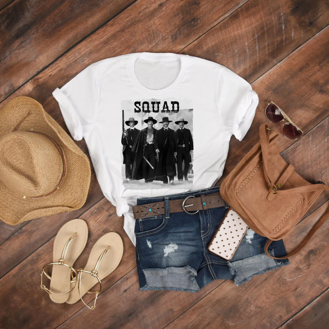 Tombstone OK Corral Squad Fan Art Sublimation Transfer T-shirt, featuring a black-and-white print of four cowboys with the word 'SQUAD' above, laid out on a wooden surface with accessories like a straw hat, sunglasses, distressed denim shorts, a tan handbag, beige sandals, and a polka dot phone case.