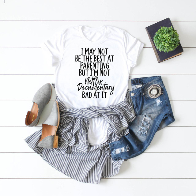 Not The Best Parent But Not Netflix Documentary Bad Sublimation Transfer T-shirt, white T-shirt with humorous quote printed on it, casual denim shorts, grey casual shoes, and a plant on a white wooden background.
