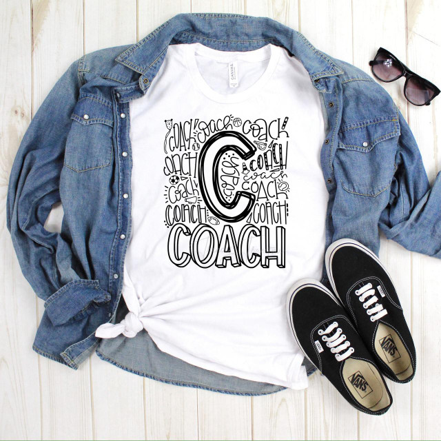 Coach Typography Sublimation Transfer, a white t-shirt with black hand-drawn style coach letter and logo design, laid flat with a denim jacket, black sunglasses, and black Vans sneakers on a wooden surface.
