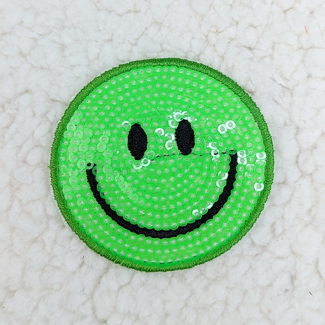 GREEN Sequin Happy Face HAT/POCKET Patch, a circular patch decorated with shiny green sequins forming a smiling face with black eyes and mouth on a white textured background.