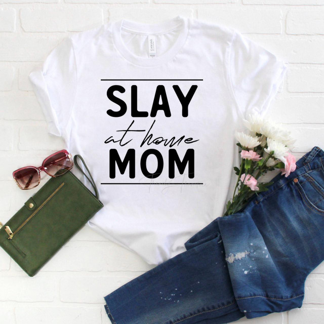 Slay at home mom sublimation transfer, white T-shirt with black and cursive font design, surrounded by sunglasses, a green wallet, pink and white flowers, and distressed blue jeans.