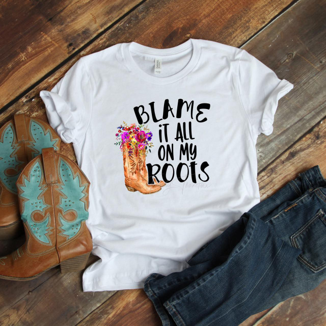 Blame it all on my roots Floral Boots Sublimation Transfer, a white t-shirt featuring a graphic of a cowboy boot filled with colorful flowers on a wooden background.