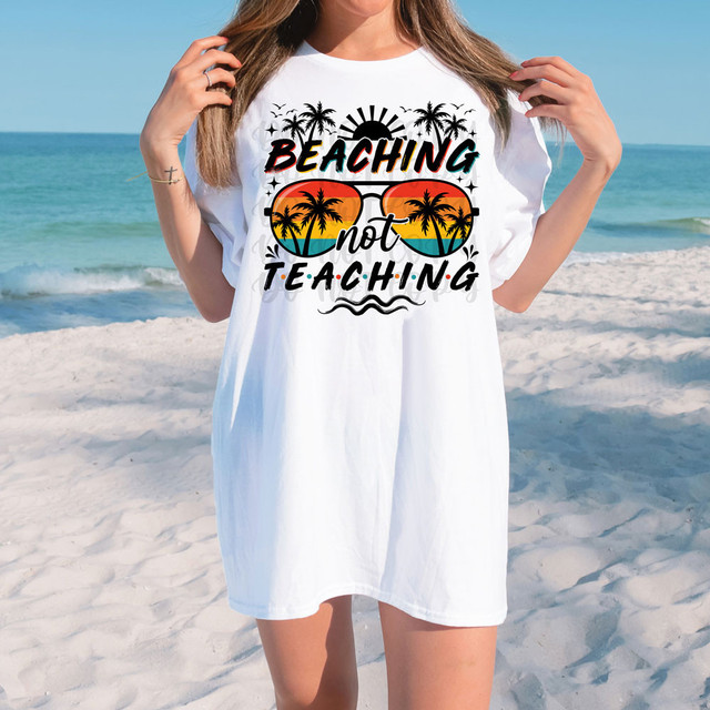 Beaching Not Teaching DTF Heat Transfer, woman wearing a white T-shirt with a beach and palm tree design on a sandy beach with ocean in the background.