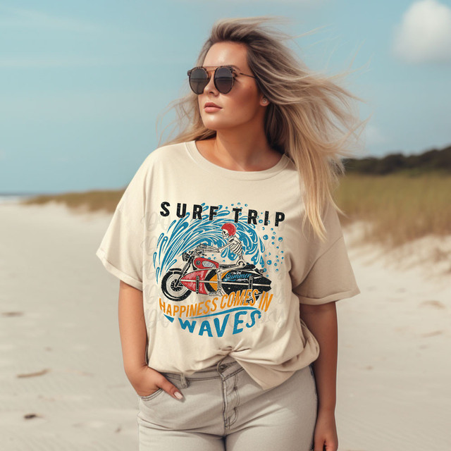 Surf Trip DTF Heat Transfer, woman wearing a beige t-shirt with a surfing and waves graphic design, at the beach with wind-blown hair and sunglasses.