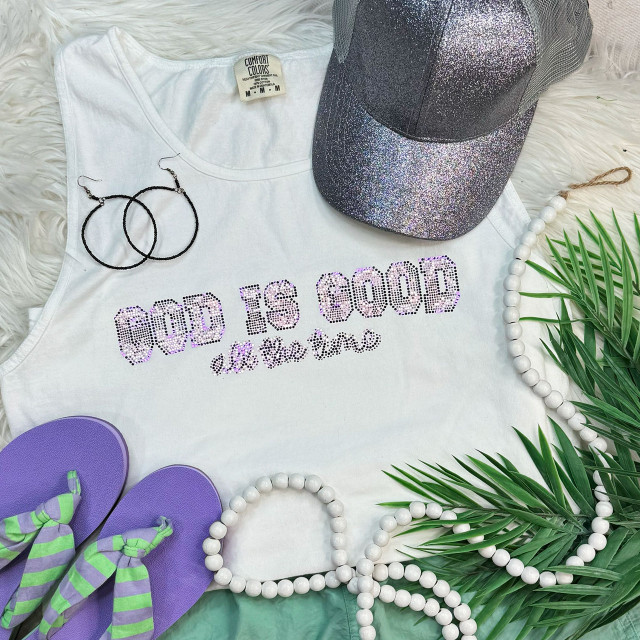 God Is Good All The Time Spangled Transfer, a decorative iron-on transfer with glittery design on a white sleeveless shirt, surrounded by a sparkly cap, purple flip-flops with green and purple striped straps, a white beaded necklace, black hoop earrings, and green palm leaves.