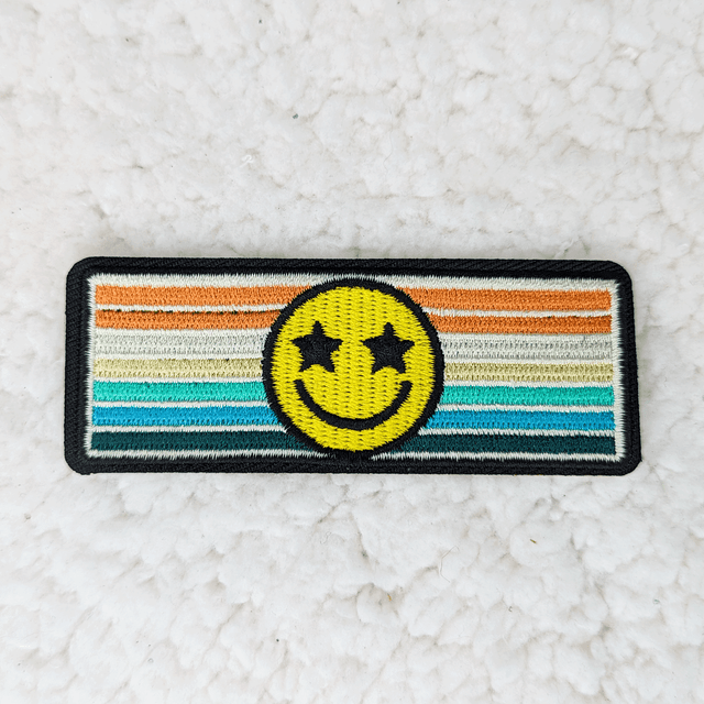 Retro Happy Face Embroidered HAT/POCKET Patch, colorful embroidered patch with a yellow smiley face having star-shaped eyes on a striped rainbow background.