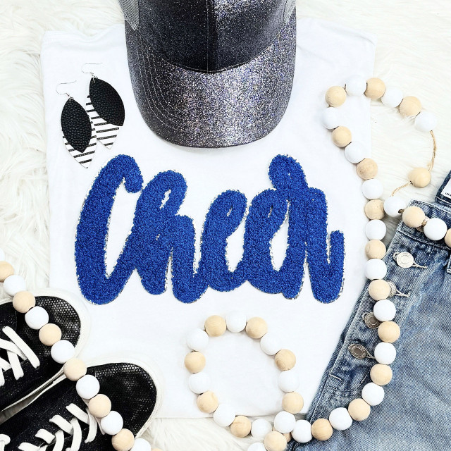 Cursive Cheer Royal Chenille Patch, vibrant blue embroidered cheer phrase on white fabric, surrounded by accessories including black sneakers, black and white feather earrings, white and natural wood bead necklace, and denim jeans.