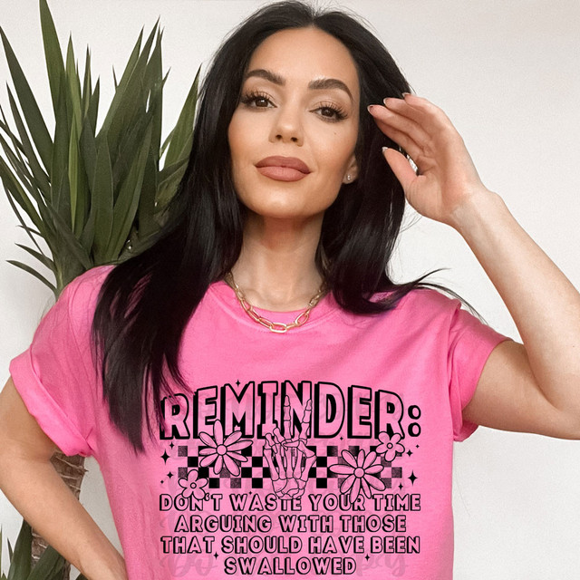 Reminder: Don't Waste Your Time Arguing With Those That Should Have Been Swallowed DTF Heat Transfer, a black and pink graphic design on a pink t-shirt worn by a woman with dark hair, with a plant in the background.