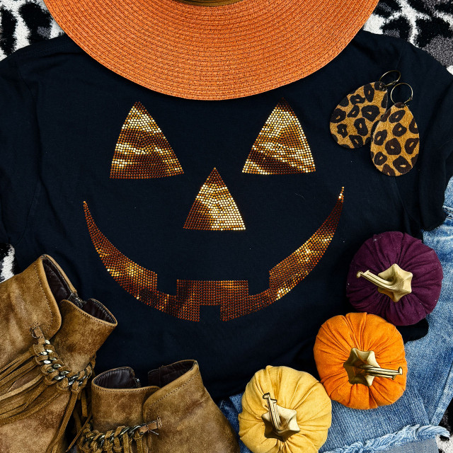 Spangled Jack O' Lantern Orange Face Heat Transfer, Halloween-themed flat lay featuring a black shirt with a spangled Jack O' Lantern face design, surrounded by orange, purple, and yellow mini pumpkins, leopard print earrings, tan boots, and an orange sun hat.