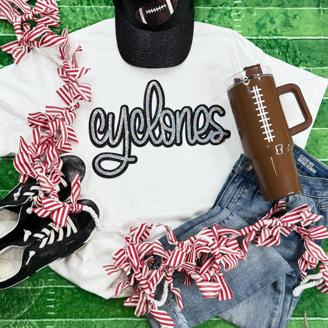 Cyclones Sequin Patch, close-up of a sports-themed sequin patch spelling 'cyclones' on a white graphic tee, surrounded by football shoes, a brown sports tumbler, and red and white striped ribbon decorations on a green surface.