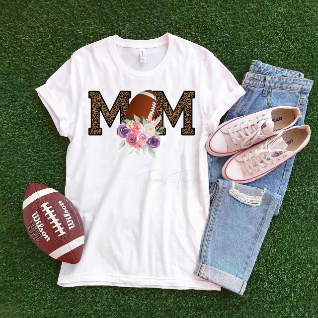 Leopard Floral Football Mom Sublimation Transfer, sports-themed T-shirt with leopard print 'MOM' text, football graphic, and floral design, displayed with sneakers, jeans, and a football on green grass.