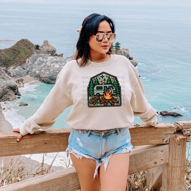 Camping Faux Patch DTF Heat Transfer, a colorful embroidered patch depicting a camping scene with a camper, trees, fire, and mountains, applied on a sweatshirt worn by a woman outdoors near the coast.