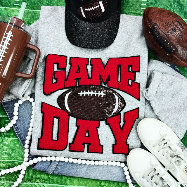 Red Chenille Game Day with Sequin Football Patch, close-up of a gray t-shirt with red lettering and a sequin football patch, surrounded by sneakers, football, drink tumbler, and accessories on a green background.
