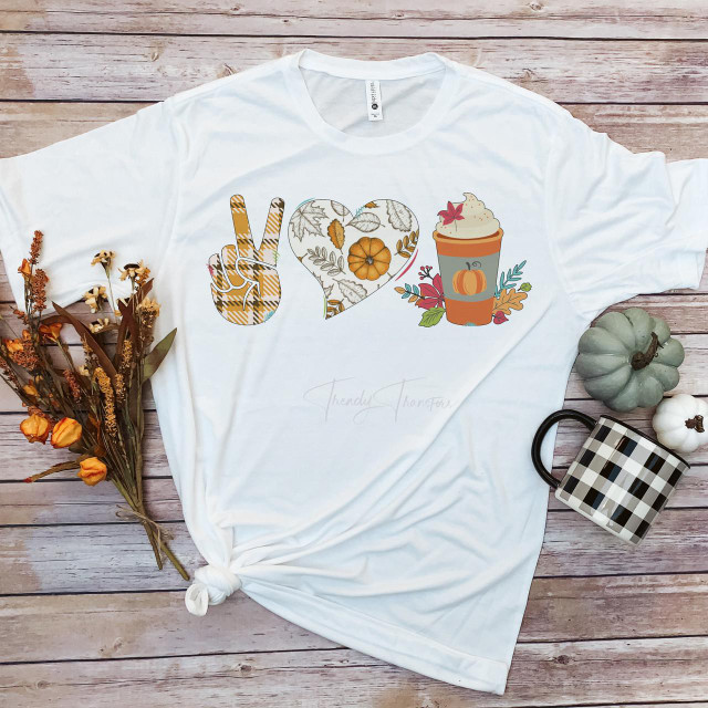 Peace Love and Pumpkin Spice Sublimation Transfer T-shirt with fall-themed design, including a peace sign, a heart with autumn leaves, and a pumpkin spice latte, laid on a wooden surface with flowers, pumpkins, and a mug around it.