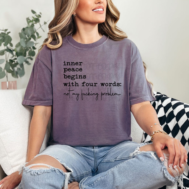 Inner Peace Begins With Four Words: Not My Fucking Problem DTF Heat Transfer, T-shirt worn by a woman sitting on a couch with distressed jeans and a cozy background.