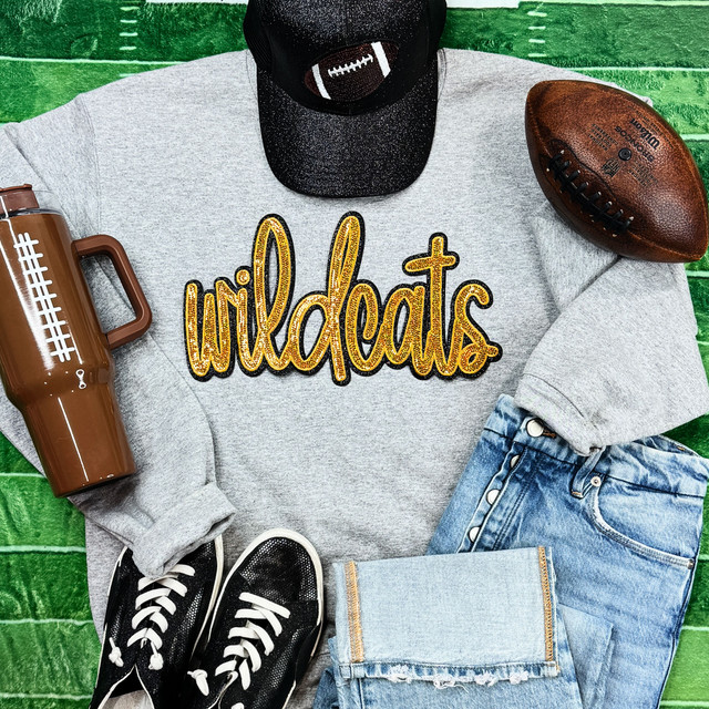 Wildcats Gold Sequin Patch, decorative patch with the word Wildcats in gold sequins placed on a casual outfit with a gray sweatshirt, jeans, and accessories like a cap, football, thermos, and sneakers on a green wooden background.