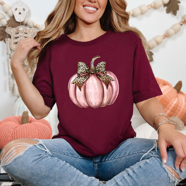 Leopard Pink Pumpkin DTF Heat Transfer, colorful pumpkin with a leopard print bow on a maroon T-shirt worn by a smiling woman.